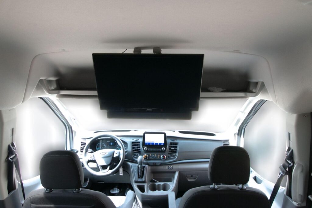 The image shows the interior of a van with a large flat-screen TV mounted on the ceiling, along with the dashboard and seats.