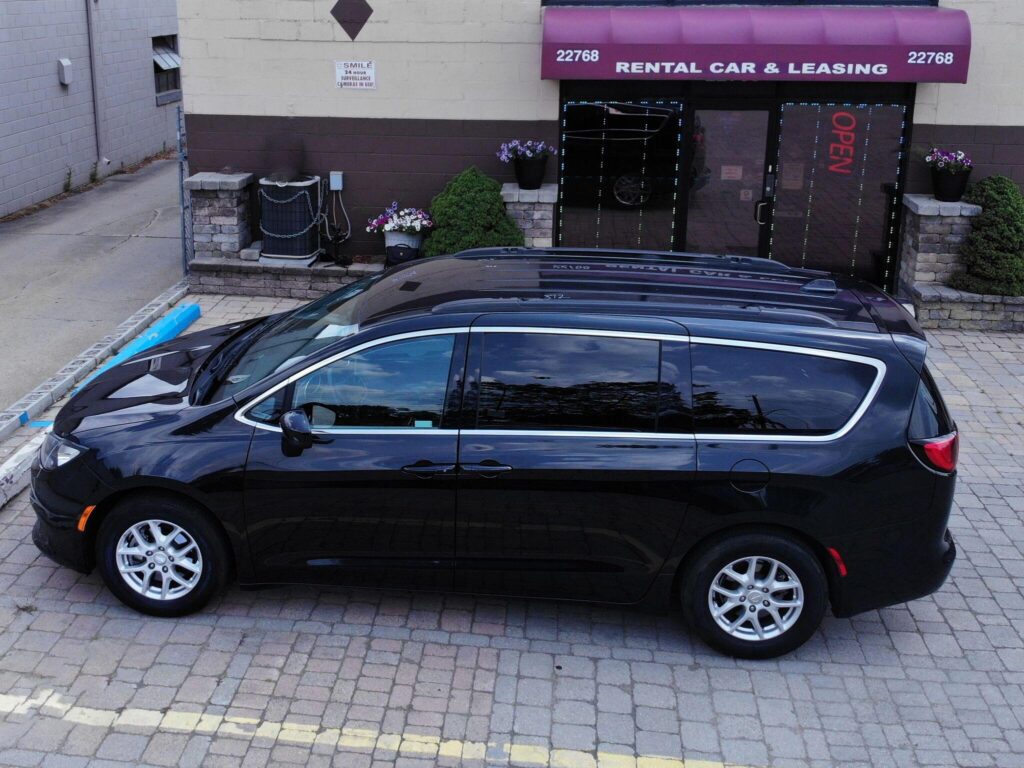 The image shows a black minivan parked in front of a rental car and leasing office
