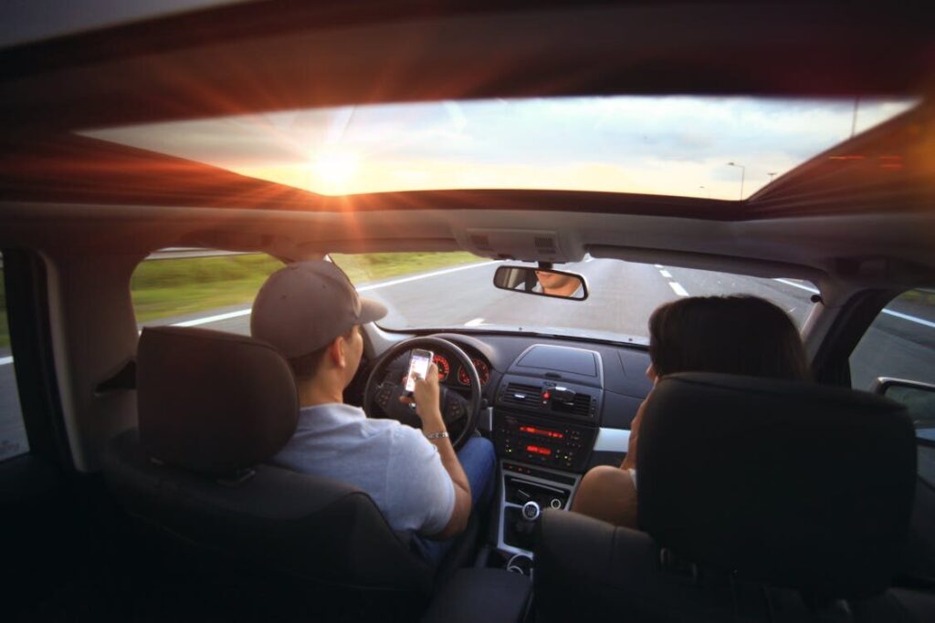 The image shows a view of a person driving a car with a passenger beside them, as the sun sets in the background