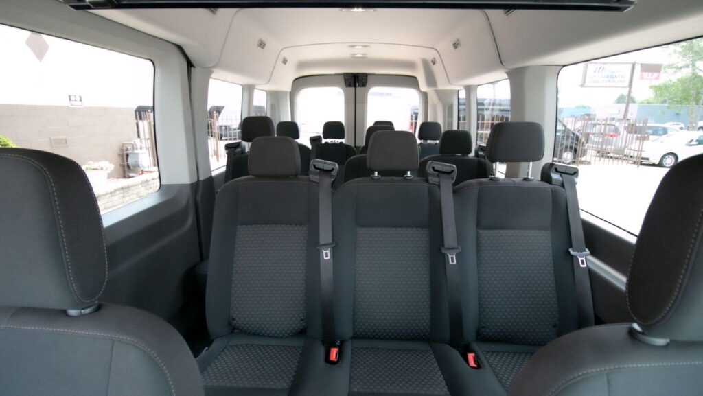The image shows the interior of a passenger van with multiple rows of gray fabric seats and seat belts